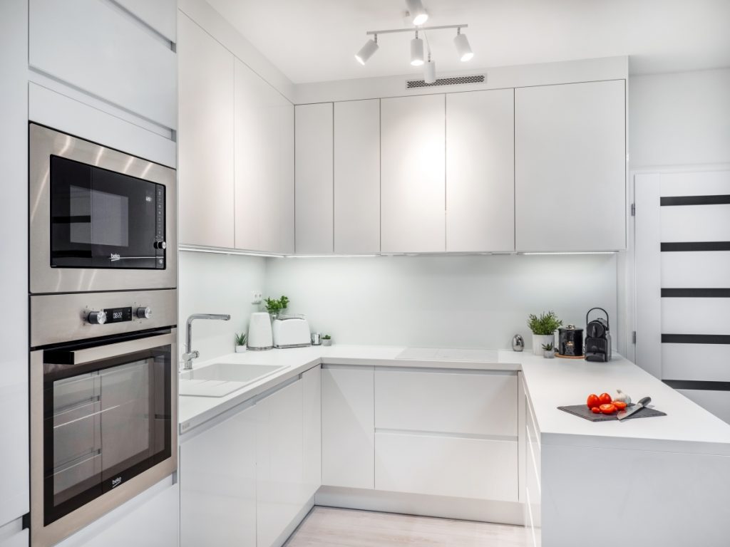 Minimalistická kuchyně v bílé barvě se stříbrnými spotřebiči. Kuchyně má tvar do U.
